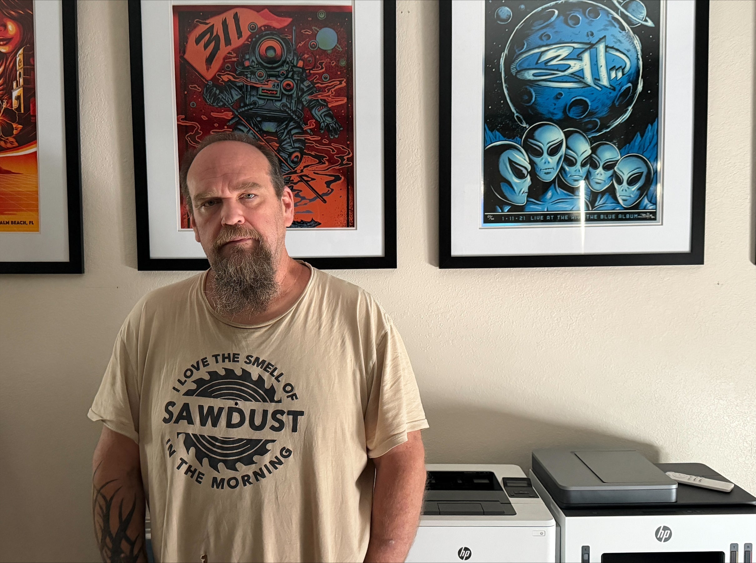 Sparky standing at home wearing a tan t-shirt reading 'I Love the Smell of Sawdust in the Morning,' with framed 311 concert posters on the wall behind him, on Day 3 of his heart health journey.